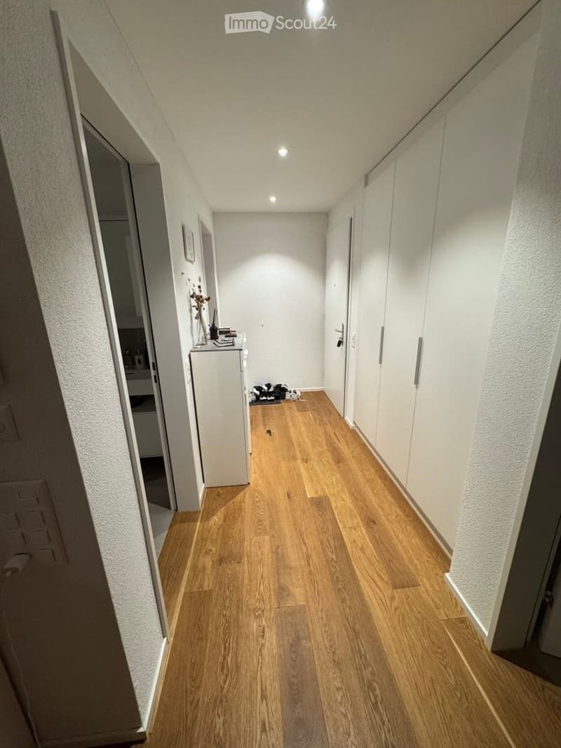 A narrow hallway with light wooden flooring, white walls, and built-in white cabinets on one side. A small table with a vase and a few items sits against the wall, and a door is visible at the end of the hallway.