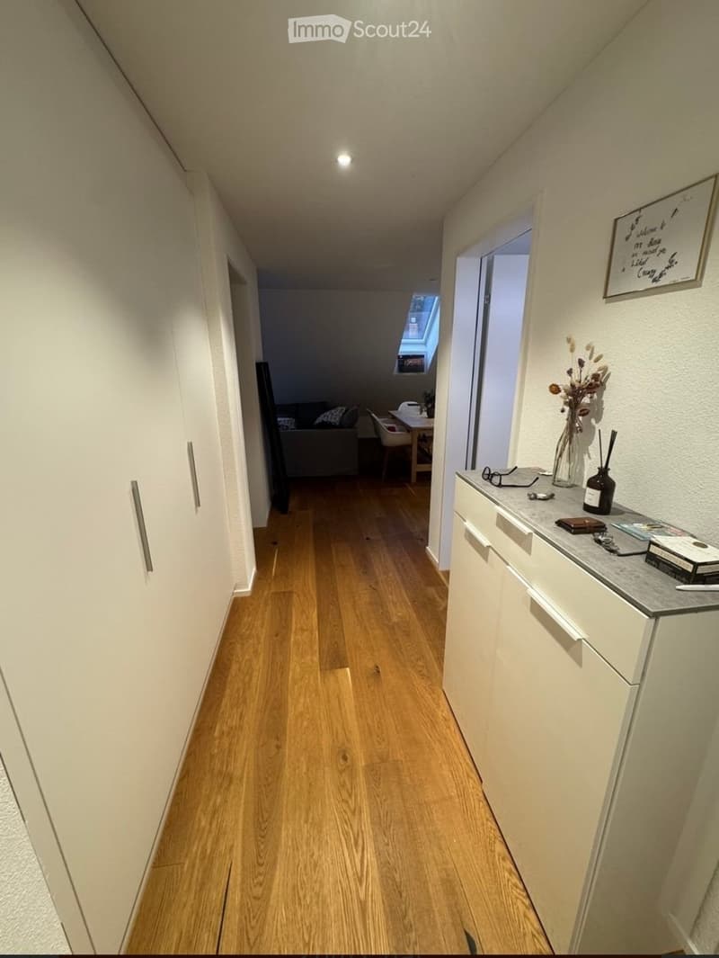 A narrow hallway with wooden flooring, white walls, and built-in storage cabinets. A small table with a vase and decorative items is visible on the right side, and a glimpse of a living area with a sofa and dining table can be seen at the end of the hallw