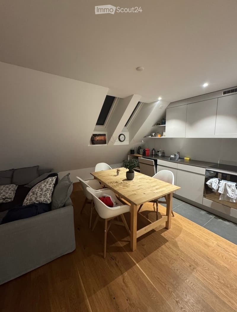 A modern, open-plan living and dining area with a grey sofa, wooden dining table, white chairs, and a sleek kitchen area featuring stainless steel appliances and cabinets.