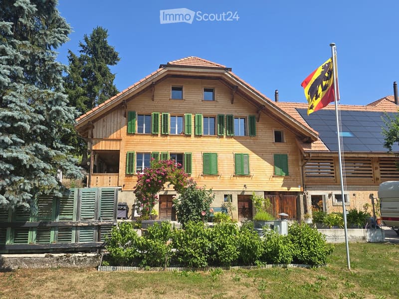 Two-story wooden house with green shutters, multiple windows, and a flagpole with a flag. The house has a well-maintained garden with various plants and trees.