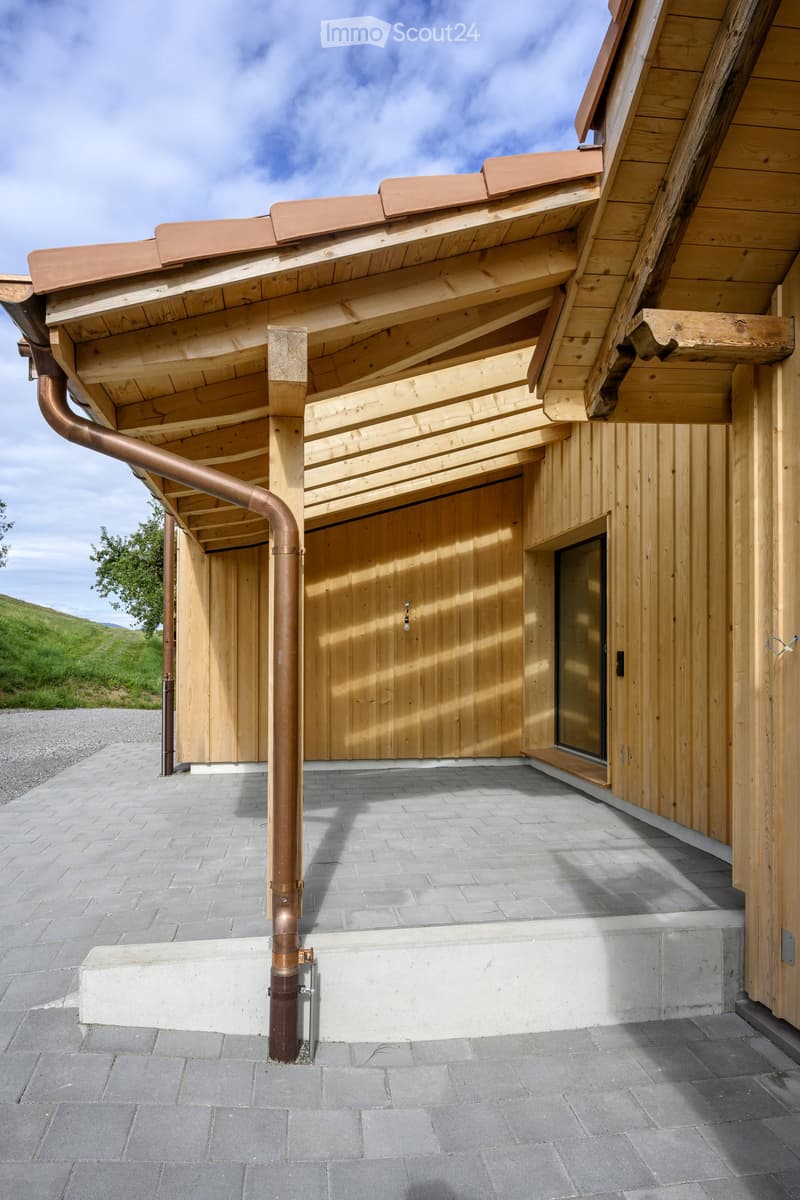Modern wooden building with a copper roof, large wooden garage door, and a glass window. The entrance features a raised concrete platform and a copper downspout.