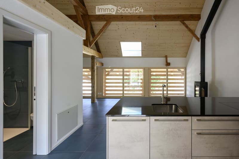 Modern kitchen with dark countertops, stainless steel sink, exposed wooden beams, and large windows with wooden shutters.