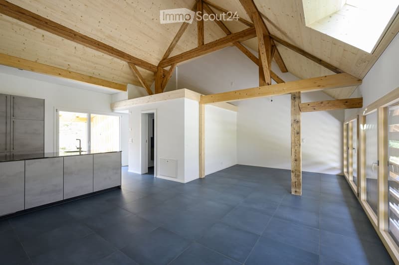 Open-plan space with exposed wooden beams, dark tiled floor, grey cabinetry, and large windows leading to an outdoor area.