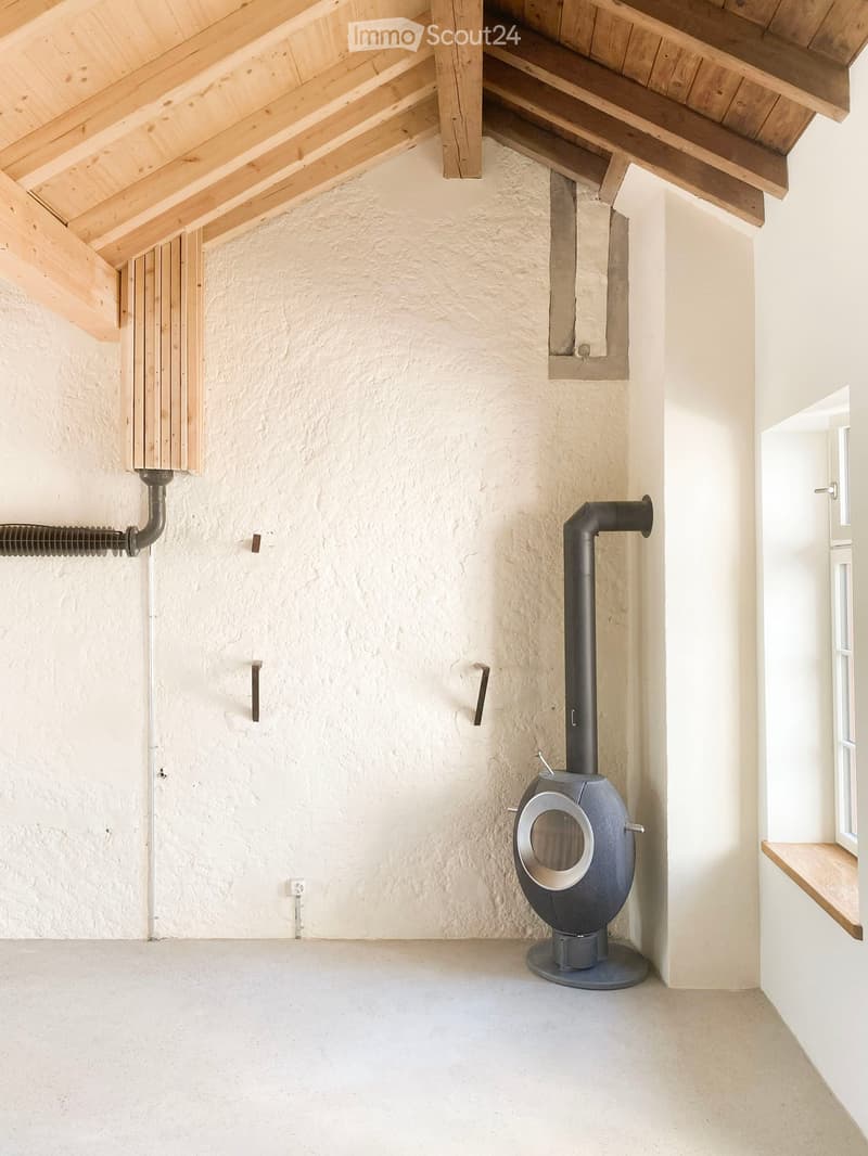 Empty room with exposed wooden beams on the ceiling, a stone wall, a modern wood-burning stove, and a small window with a wooden sill.