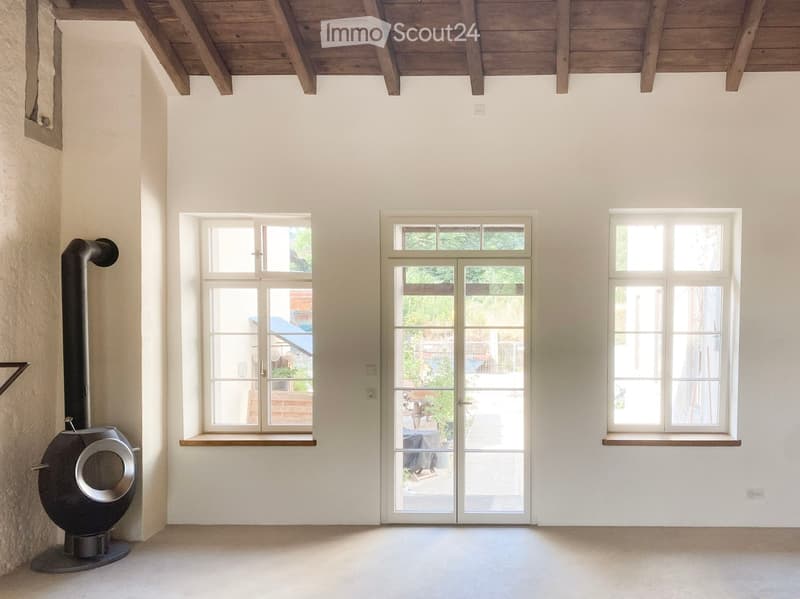 Empty room with white walls, wooden ceiling beams, large windows, and a modern wood-burning stove with a chimney pipe.