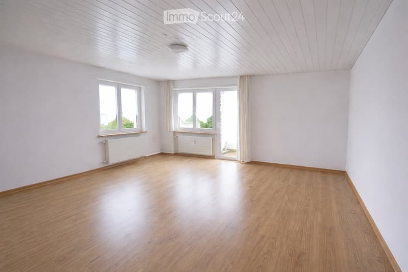Empty room with wooden flooring, white walls, and large windows with curtains. Radiators are visible along the walls.