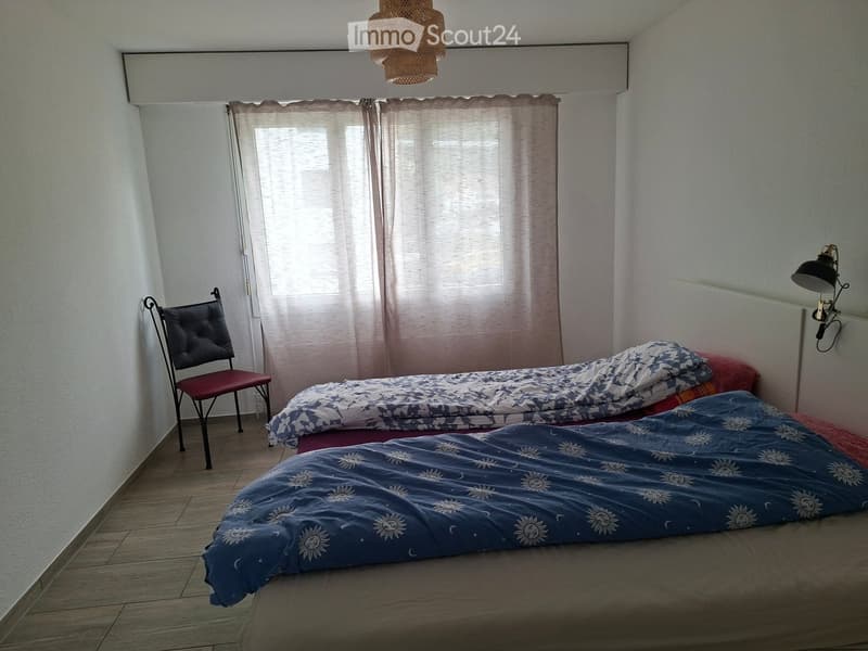 Bedroom with two single beds, a chair, and a wall-mounted lamp. Light-colored walls and wooden flooring.