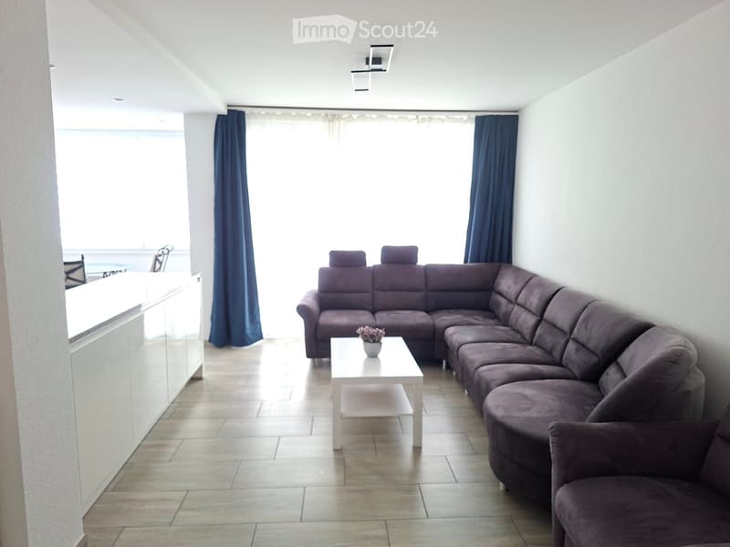 Modern living room with a large sectional sofa, white coffee table with a small flower pot, and a dining area with chairs and a table visible through an open doorway.