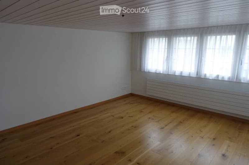 Empty room with wooden flooring, white walls, and large windows with white curtains. No furniture or equipment present.