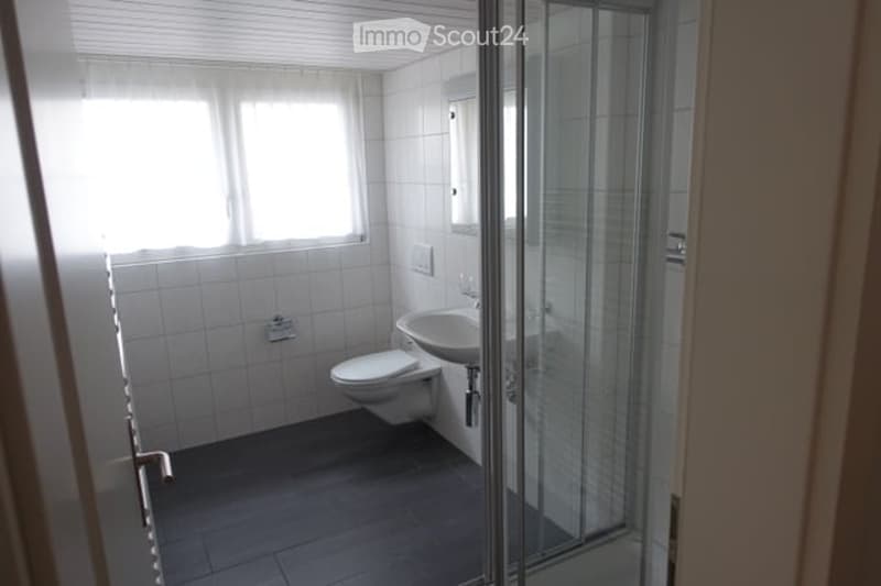 Modern bathroom with white tiles, a toilet, a sink, a mirror, and a glass shower enclosure. The room is well-lit with a window providing natural light.