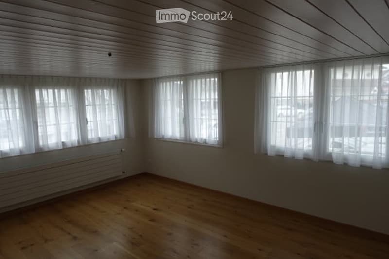 Completely empty room with wooden flooring, white walls, and large windows with white curtains. No furniture or equipment present.