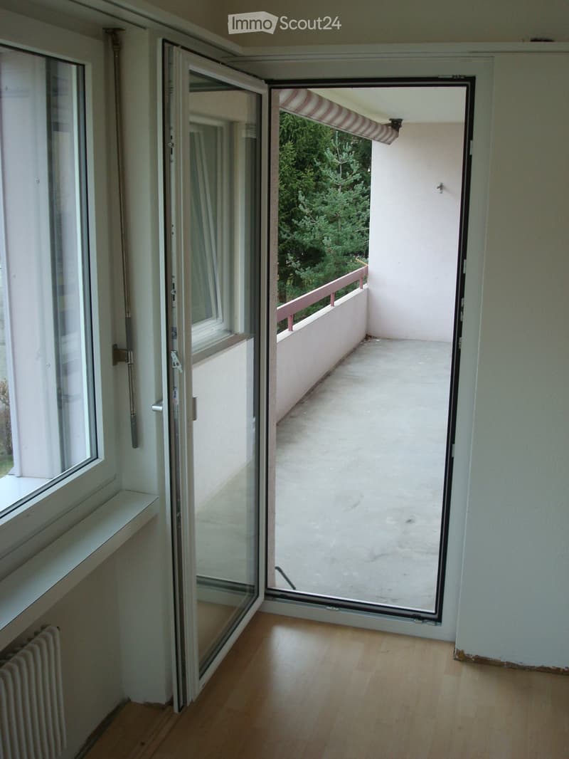 Open sliding glass door leads to a small balcony with a wooden railing and striped awning, surrounded by greenery.