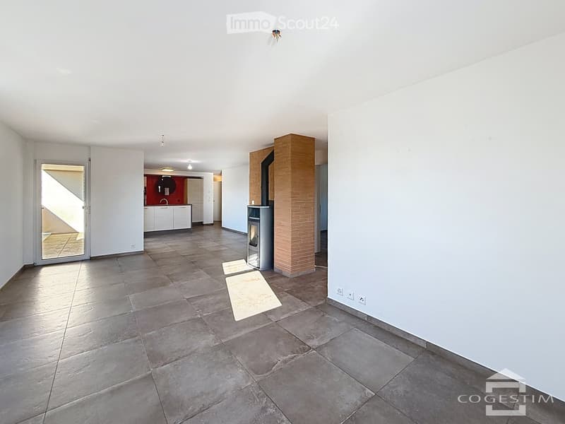 Empty room with tiled floor, white walls, and a brick pillar. A small kitchen area is visible in the background, but no furniture is present.