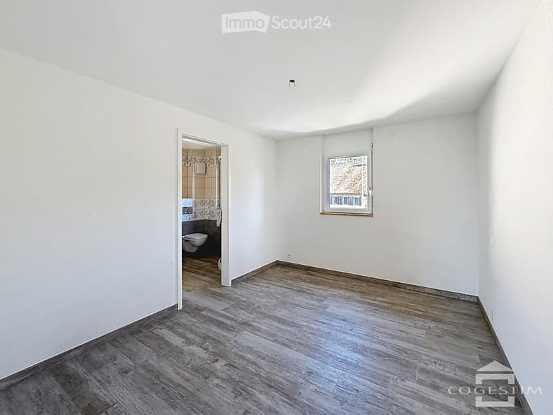 Empty room with wooden flooring, white walls, and a window. An adjacent bathroom is visible through an open doorway.