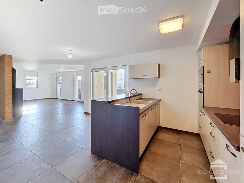 Modern kitchen with light wooden cabinets, stainless steel sink, and a central island. The space features tiled flooring and large windows with sliding glass doors leading to an outdoor area.