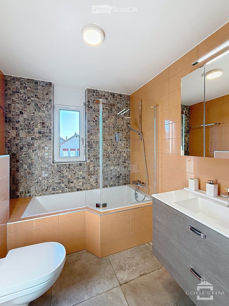 Modern bathroom with a bathtub, toilet, sink, and shower area. Features include a large mirror, modern lighting, and a window with a view.