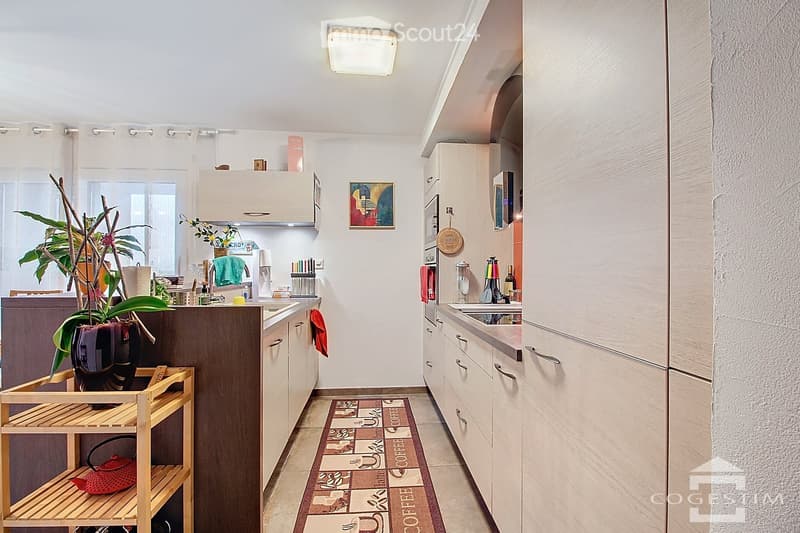 Modern kitchen with white cabinets, stainless steel appliances, and a coffee-themed rug. Features a wooden shelf with a teapot and plant, and a small wooden cart with additional items.