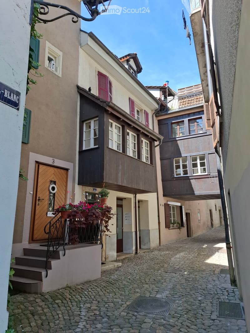 A narrow cobblestone street lined with multi-story buildings featuring wooden balconies, shutters, and potted plants. The street is flanked by traditional European architecture with varied facades and a clear blue sky above.