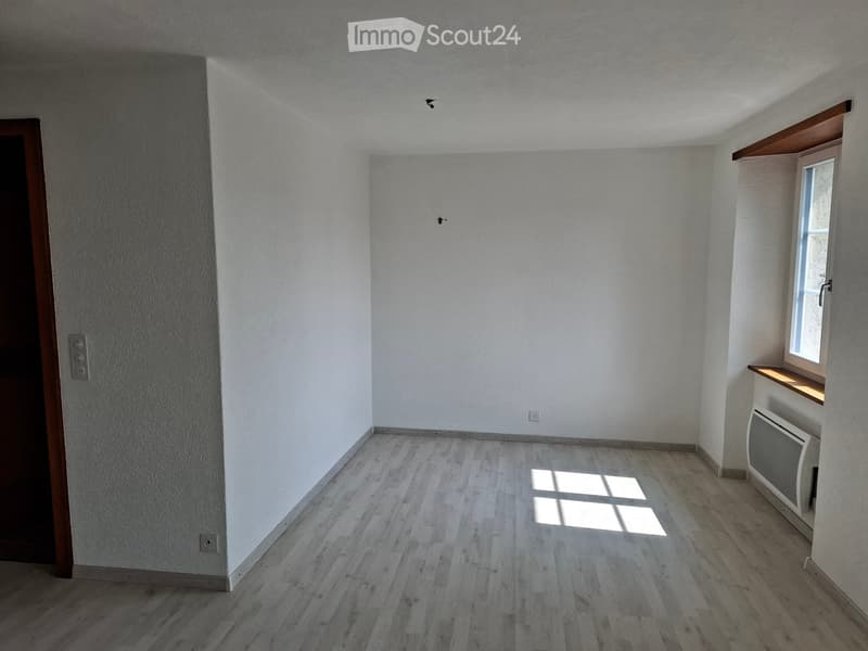 Empty room with light wooden flooring, white walls, a window with a small wooden sill, and a radiator. The room has a door on the left side and electrical outlets visible on the walls.