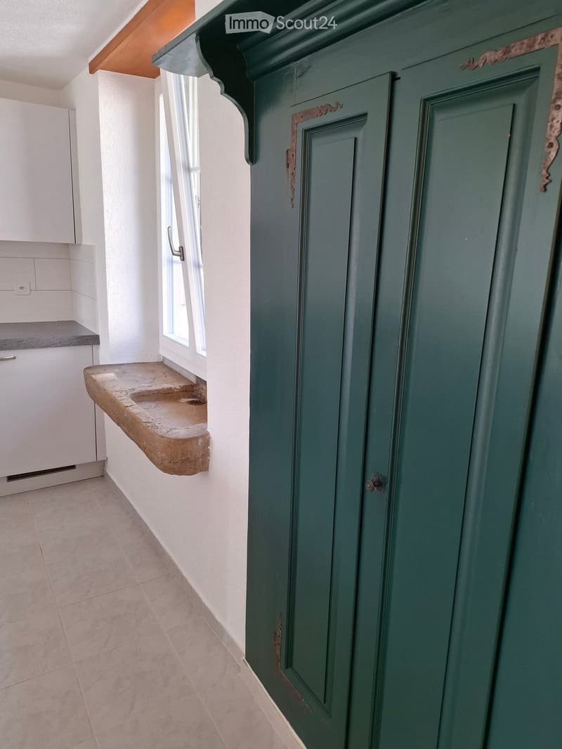 A narrow hallway with a green door on the right, a window with a stone sill on the left, and white tiled flooring.