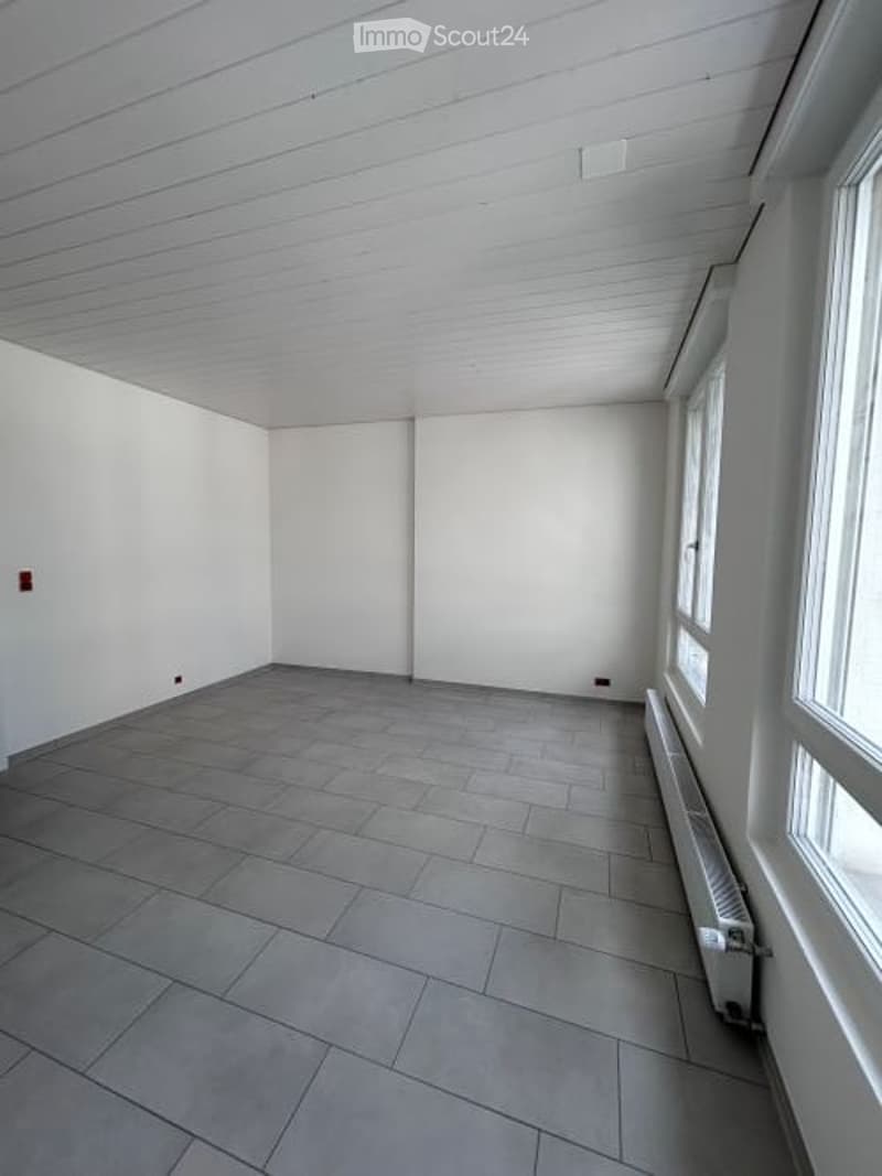 Completely empty room with white walls, grey tiled floor, and large windows on one side. No furniture or equipment present.