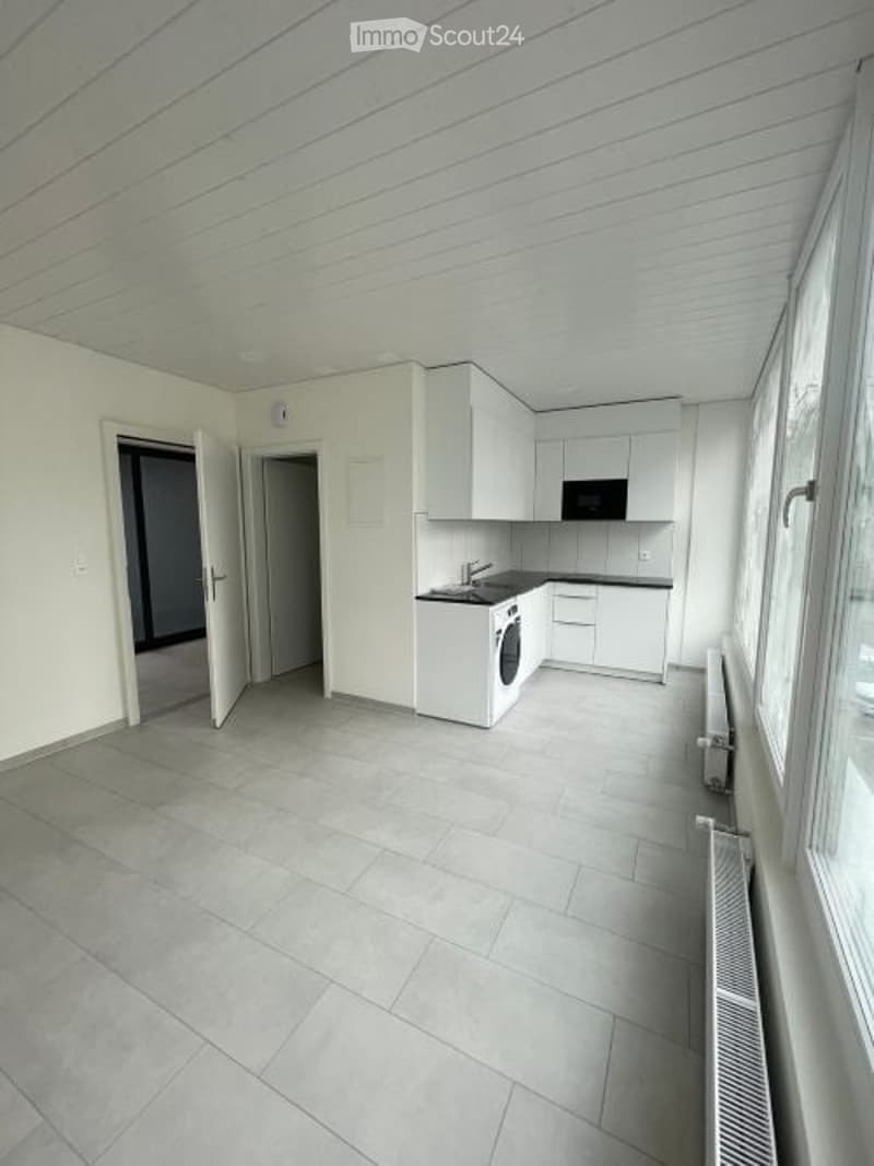 Empty kitchen with white cabinets, tiled floor, and large windows. No furniture or appliances are present.