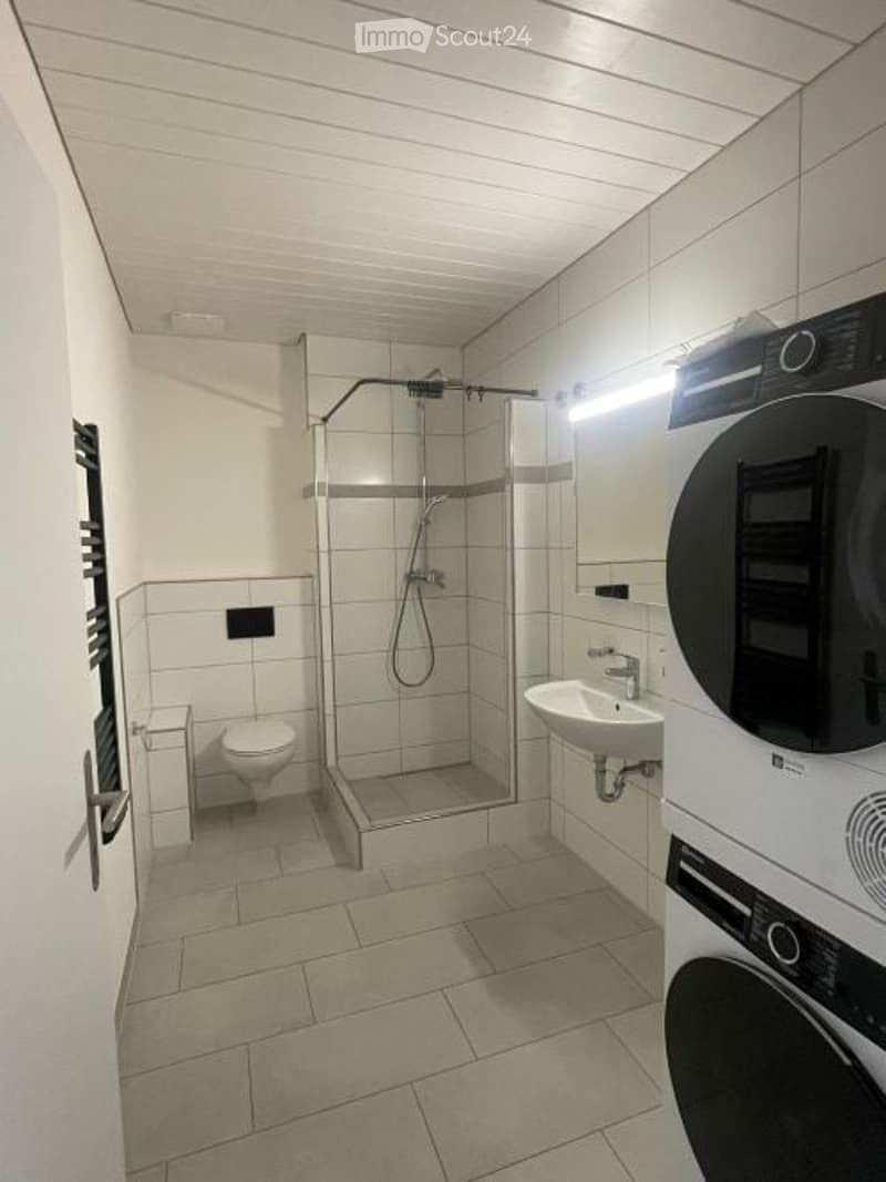 Modern bathroom with white tiled walls and floor, featuring a shower stall, toilet, sink, and a private washing machine.