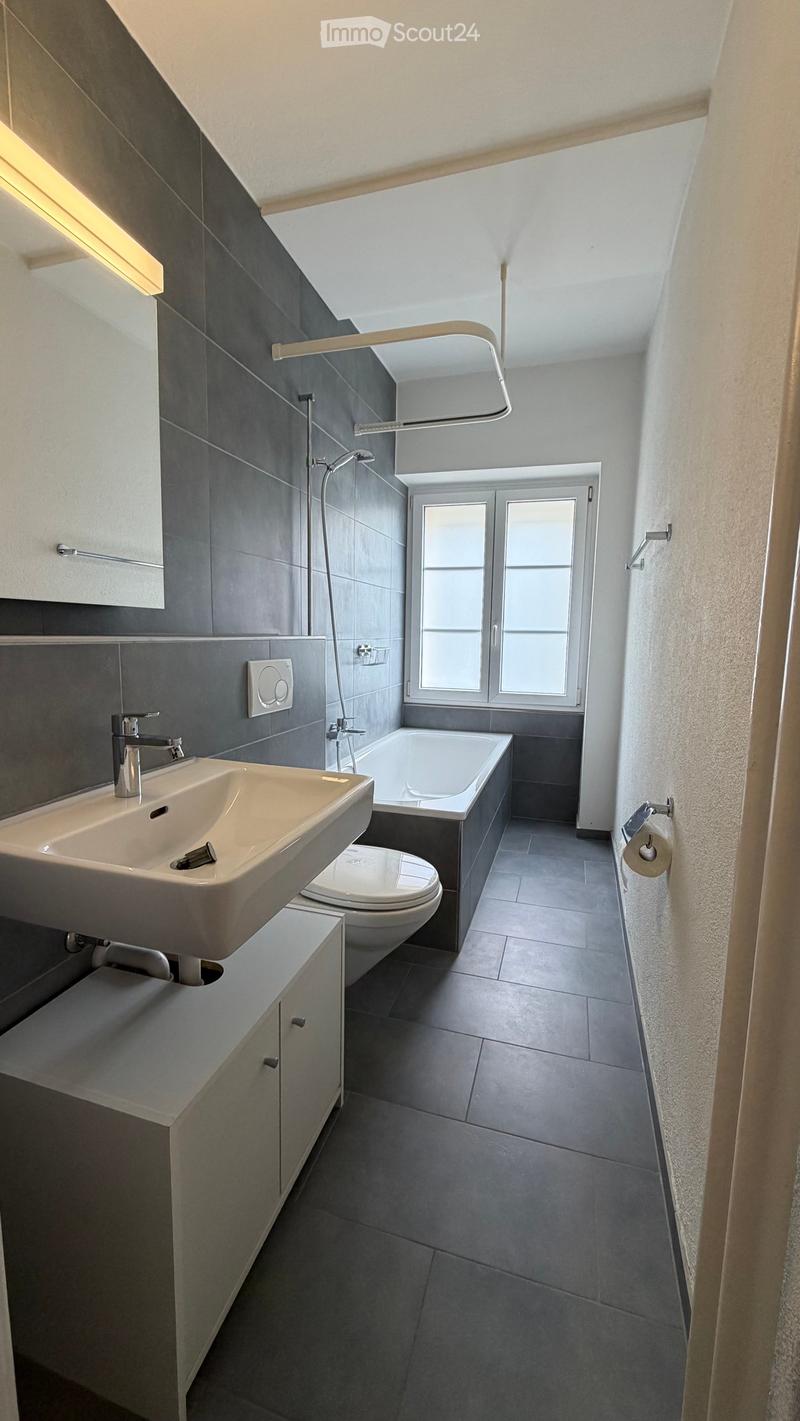Modern bathroom with grey tiles, a sink, toilet, bathtub, and shower. Features include a window, built-in cabinetry, and sleek fixtures.