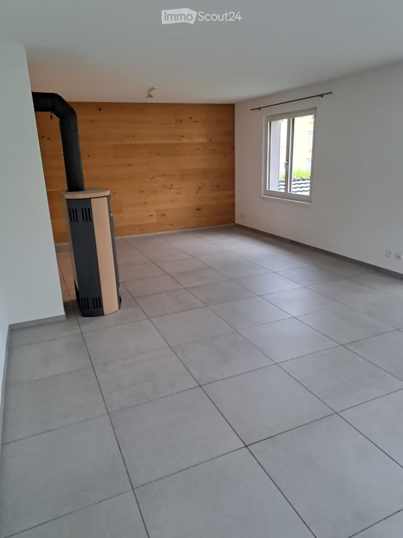 Empty room with tiled floor, white walls, and a wood-paneled accent wall. A wood-burning stove is positioned against the wall, and a window with blinds allows natural light to enter.