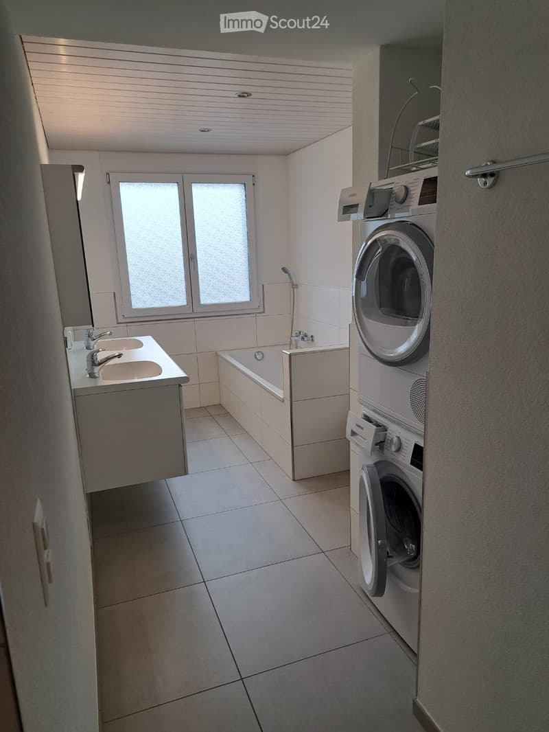 Modern bathroom with a double sink, bathtub, and stacked washer/dryer unit. Light-colored tiles and a large window provide natural light.
