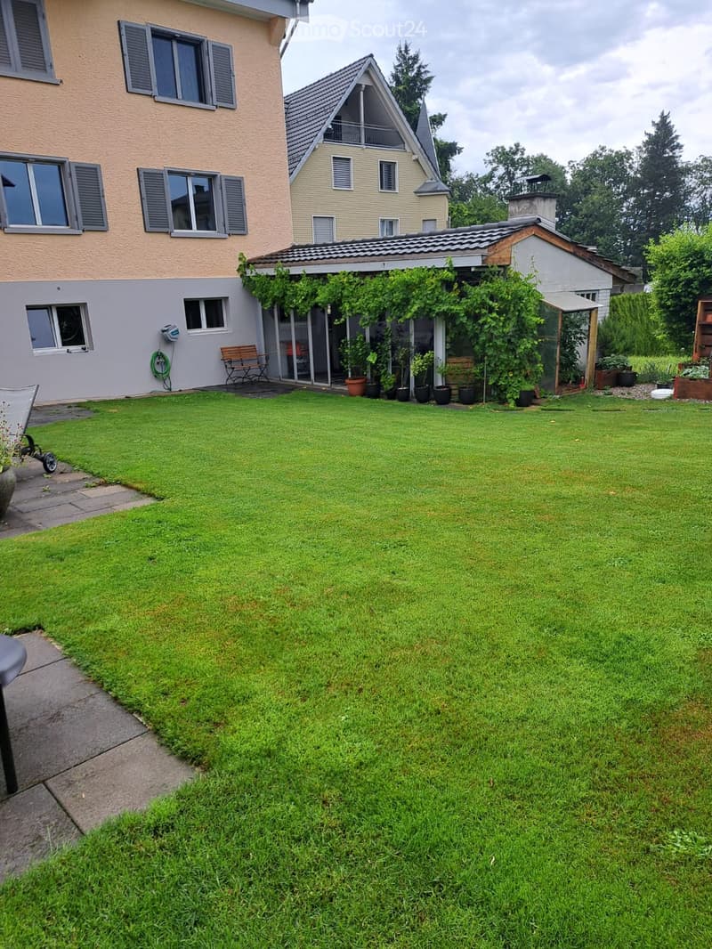 Well-maintained lawn with surrounding garden beds, potted plants, and a small seating area near the house entrance.