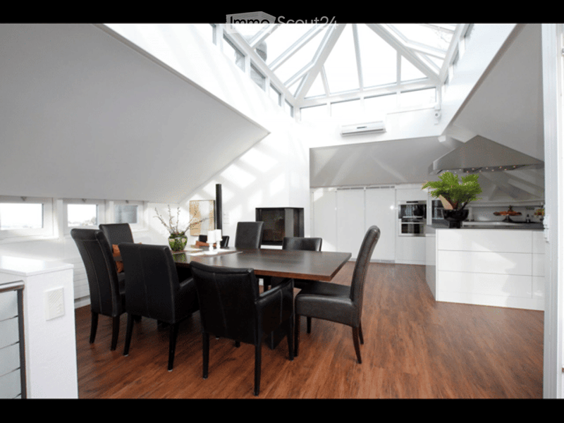 Modern, open-plan dining and kitchen area with wooden flooring, sleek white cabinets, a dining table with black chairs, and a potted plant on the counter.