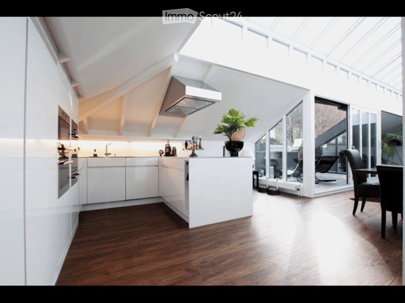 Modern kitchen with white cabinets, stainless steel appliances, wooden flooring, and a central island with a potted plant. Large windows allow natural light to fill the space.