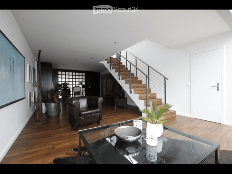 Modern living room with wooden flooring, a staircase leading to an upper level, a black leather sofa, a glass coffee table with a bowl and vase, and decorative plants.