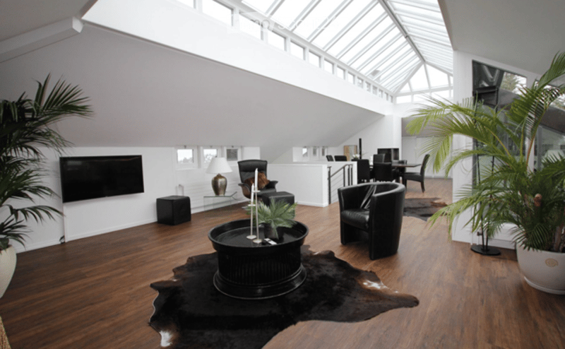 Modern living room with hardwood floors, large windows, a black leather sofa, coffee table, dining area with chairs and table, and decorative plants.