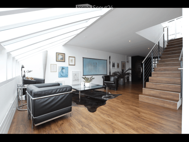 Modern living room with wooden flooring, large windows, black leather sofa, coffee table, armchair, floor lamp, and artwork on the walls.