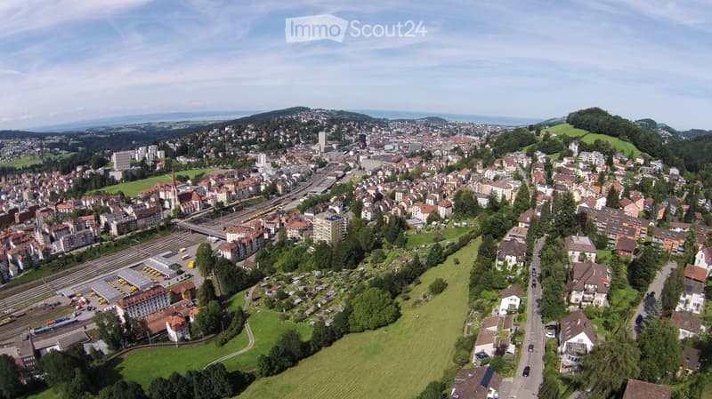 Aerial view of a town with a mix of residential and commercial buildings, surrounded by green spaces and a railway line running through the center.