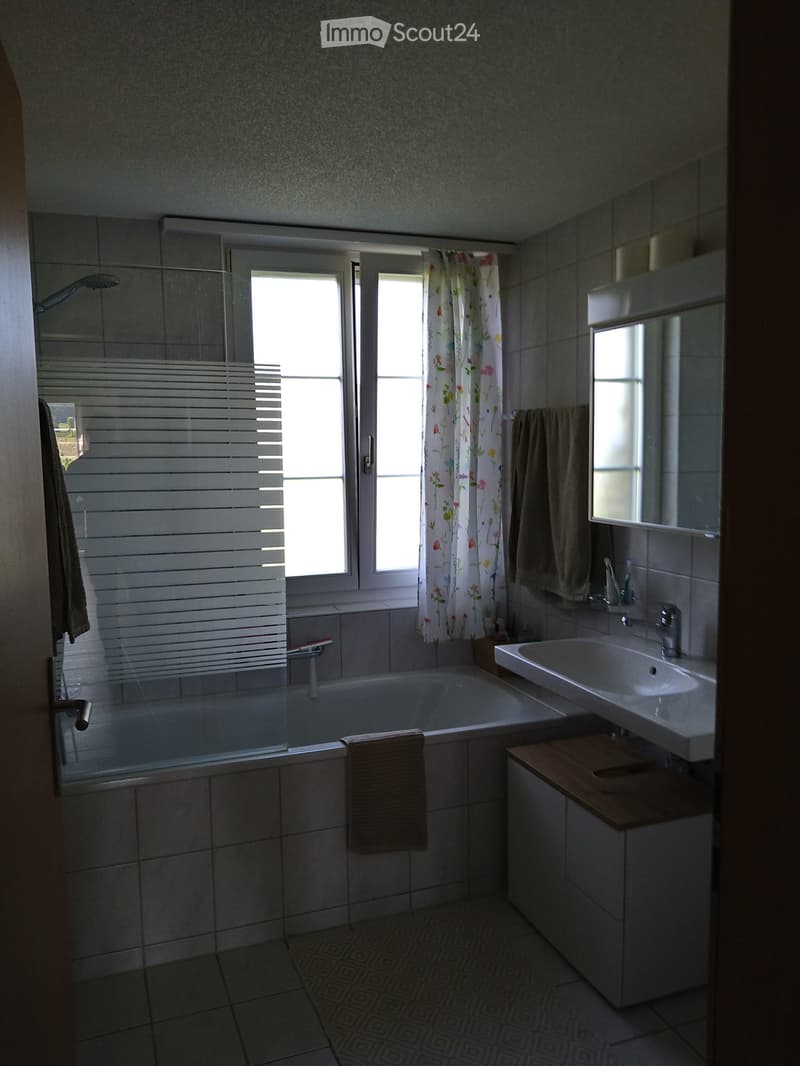 Bathroom with a bathtub, sink, mirror, and towels. Features a window with blinds and a floral curtain.