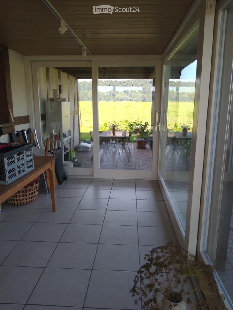 Bright, tiled balcony with large glass doors opening to a scenic view of a green field and trees. Includes a small table and chairs, potted plants, and a decorative dried flower arrangement.