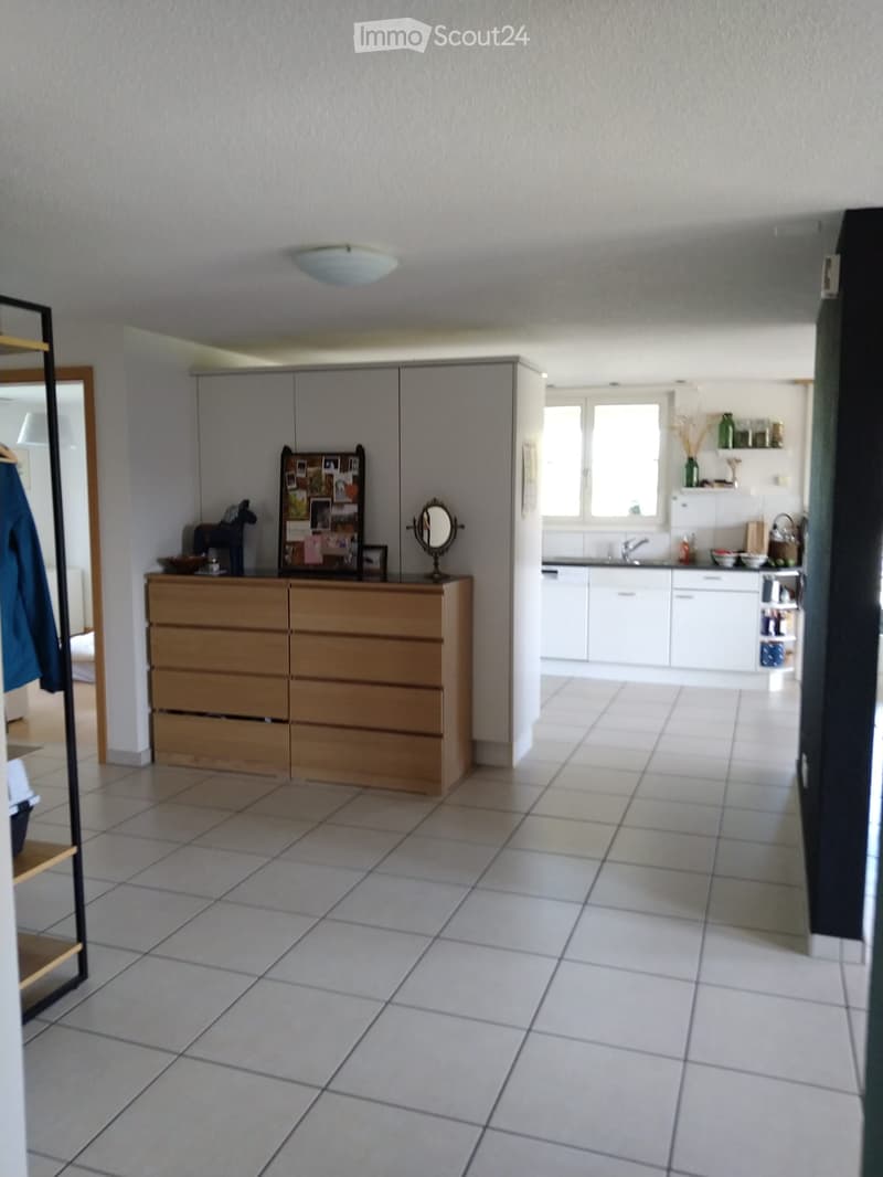 A well-lit hallway with tiled flooring, leading to a kitchen area with white cabinets, a sink, and a refrigerator. A wooden cabinet with drawers and a mirror is visible, along with a shelf holding framed photos and a vase.