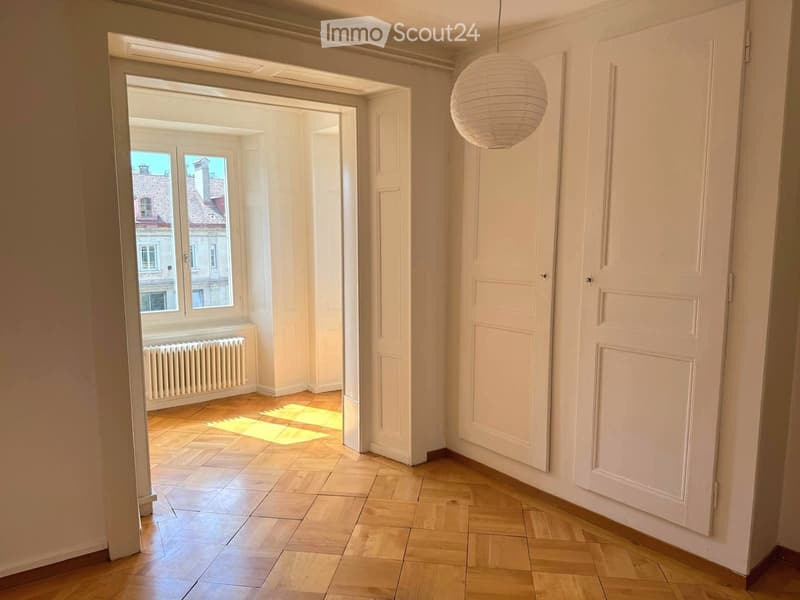 Empty hallway with wooden flooring, white walls, and a hanging spherical light fixture. A doorway leads to a room with large windows and a radiator.