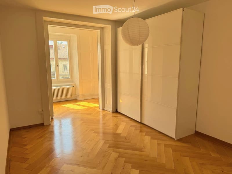 Empty hallway with wooden flooring, white walls, a hanging spherical light fixture, and an open doorway leading to another room with a window and radiator.
