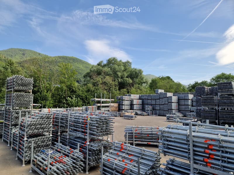 An outdoor area filled with stacks of metal pipes and construction materials, surrounded by trees and a clear blue sky.