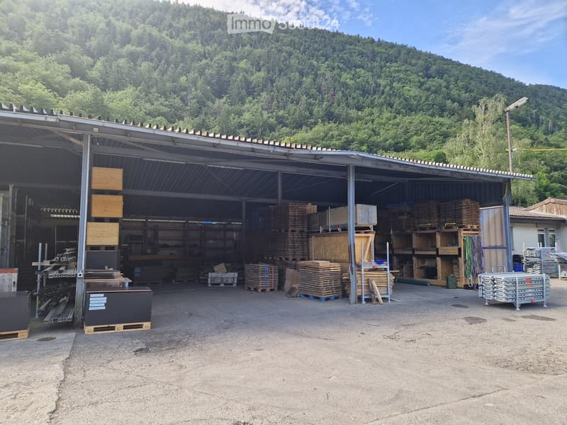 A large open-sided garage or workshop with a metal roof, filled with stacks of wooden planks, shelves, and various construction materials. The background shows a forested mountain landscape.