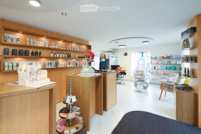 A well-lit, modern beauty salon with wooden shelves displaying various beauty products, a reception desk, a seating area with a patterned chair, and a mirror. The space features a clean, organized layout with a variety of skincare and haircare items on di