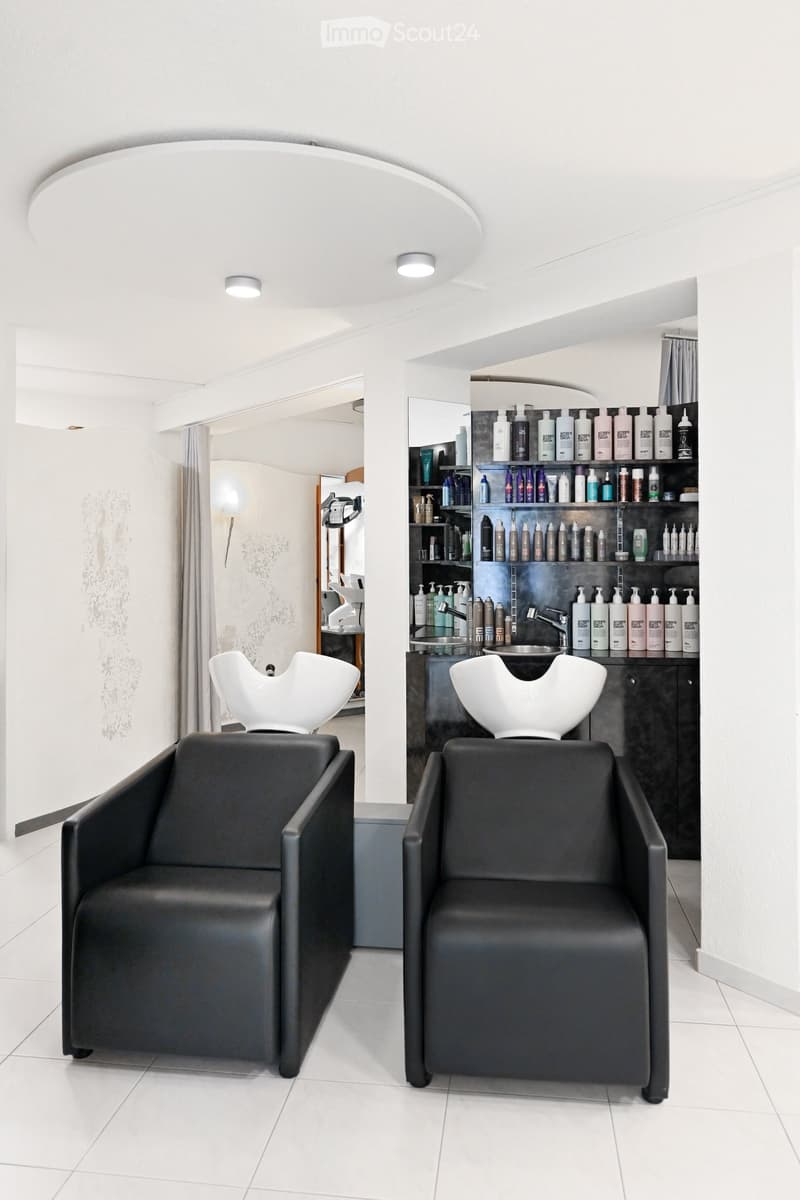 Modern salon with two black leather chairs, white sinks, shelves filled with hair products, and a mirror.