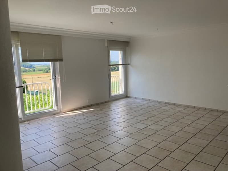 Empty room with tiled floor, large windows with blinds, and a view of a green landscape outside.