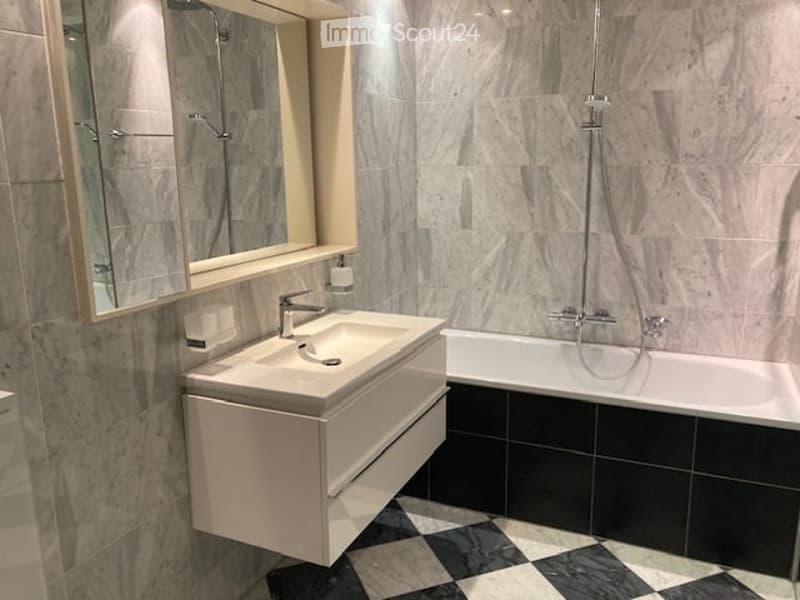 Modern bathroom with a white sink, bathtub, and large mirror. The walls are covered in marble tiles, and the floor features a black and white checkered pattern.