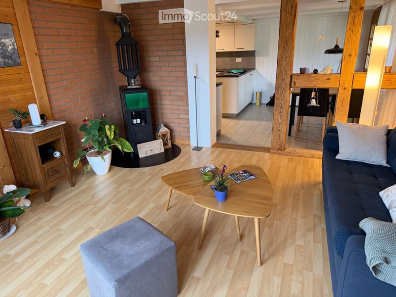 Living room with wooden flooring, a blue sofa, a wooden coffee table, a grey ottoman, a wooden cabinet, a fireplace, and a small kitchen area visible in the background.