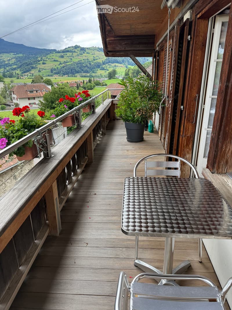 Wooden balcony with metal table and chair, potted plants, and scenic countryside view.